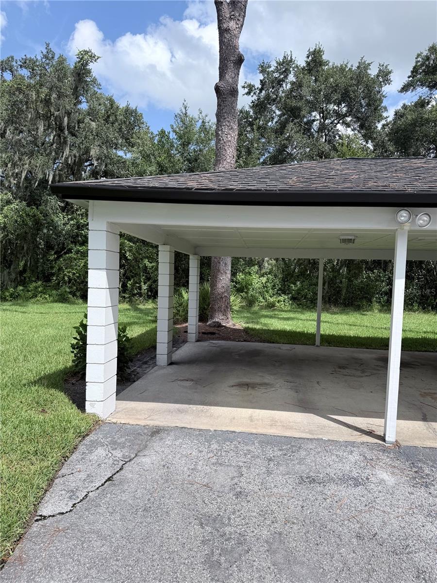 Covered carport with white supports and a concrete parking area beside a grassy, wooded yard.