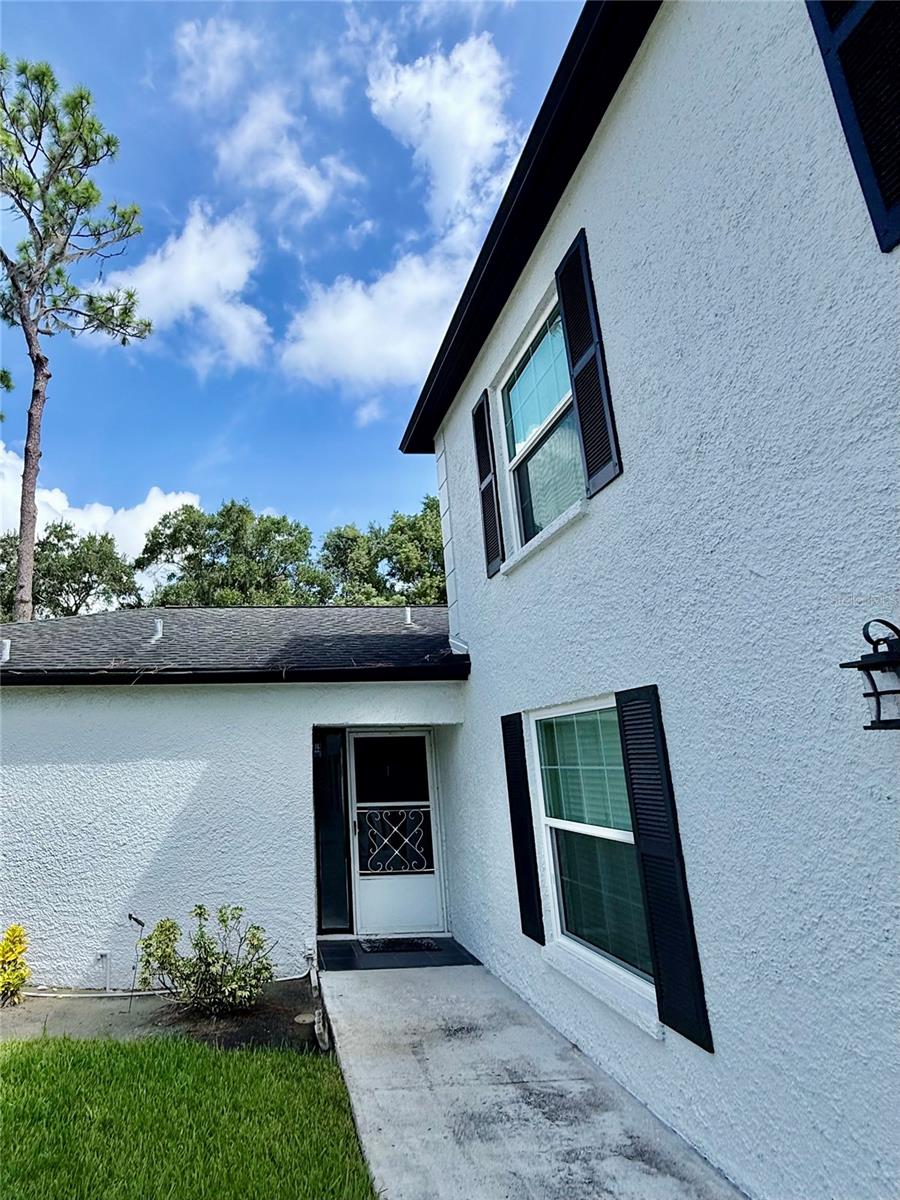 White stucco home entrance with black shutters, a concrete walkway, and front lawn.