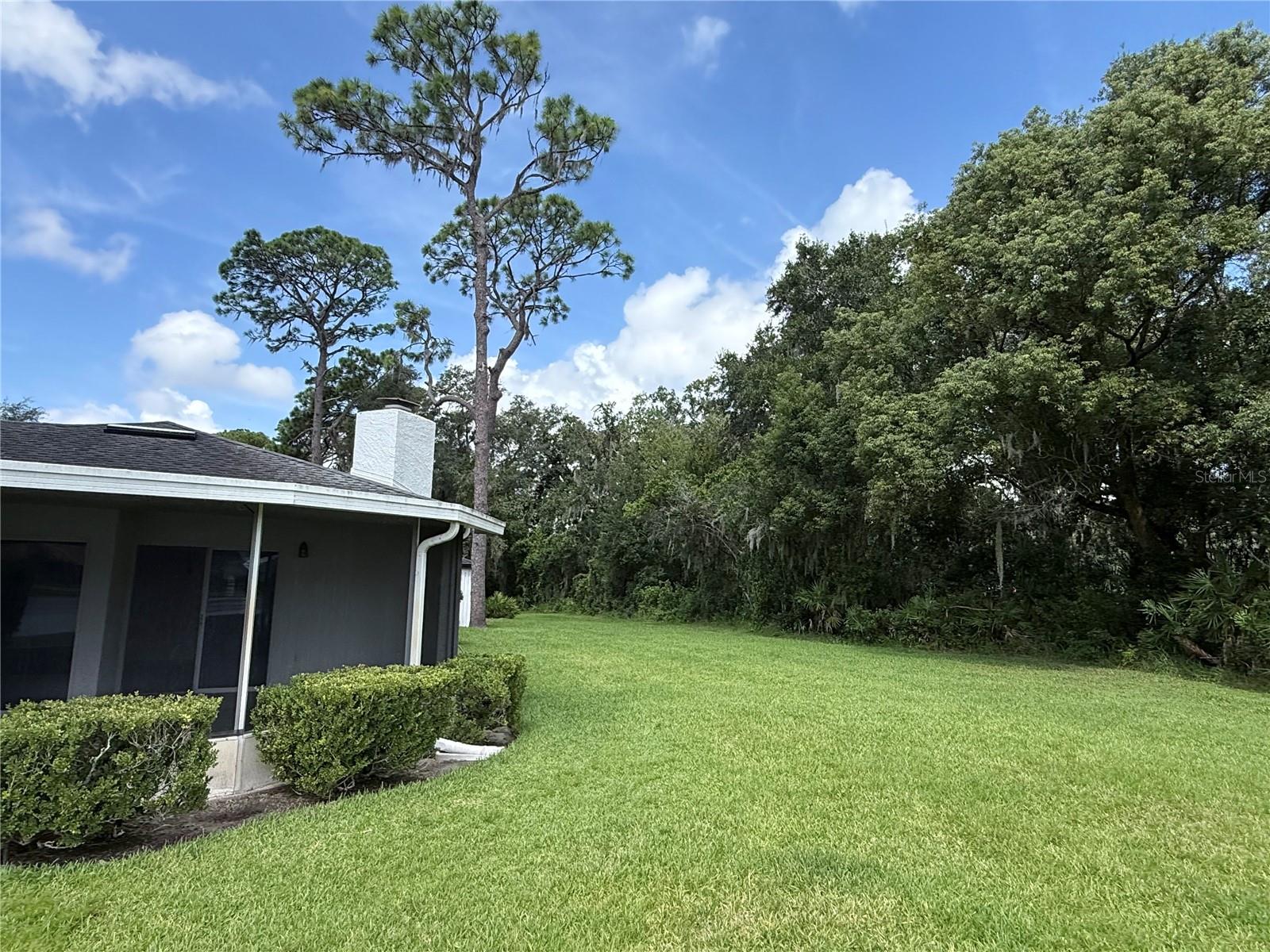 Home’s side exterior beside a broad lawn, trimmed shrubs, and a mature tree line.