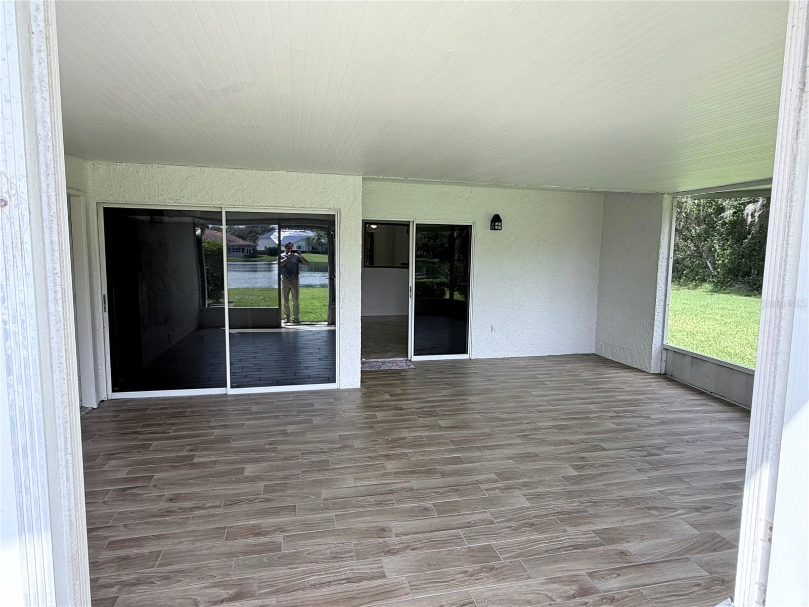 Covered lanai with wood-look tile flooring, sliding glass doors, and screened openings facing a green lawn.