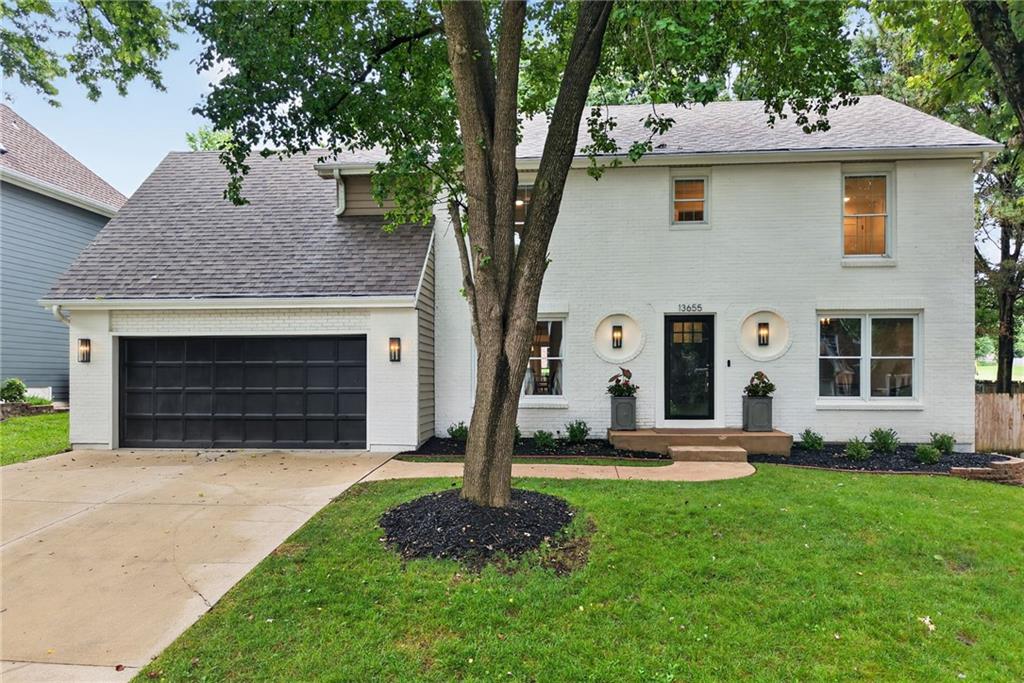 White two-story brick house with an attached dark garage and a large tree in the front lawn.