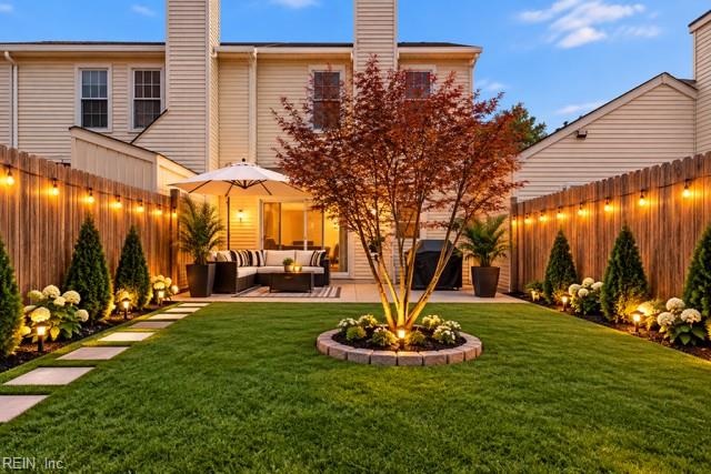 Landscaped backyard with a central tree, lawn, and a patio seating area enclosed by lit privacy fences.