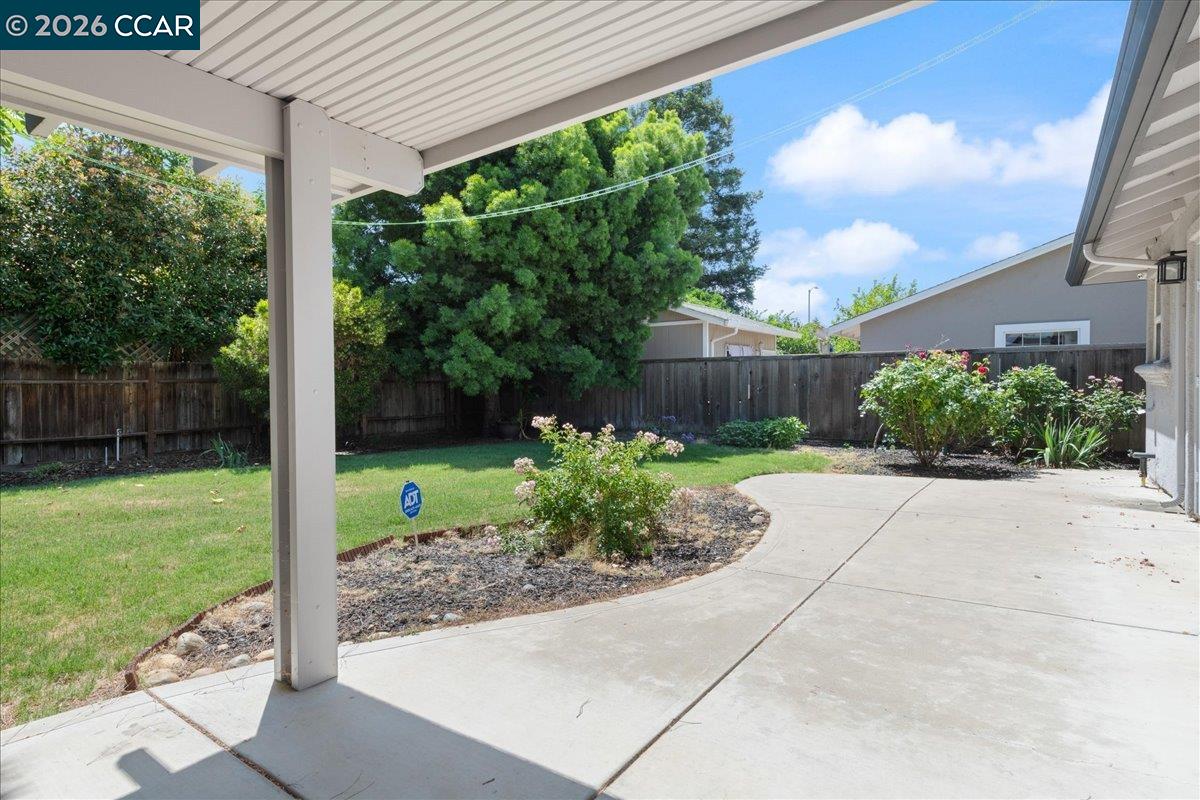 Covered patio overlooking a fenced backyard with lawn, flowering shrubs, and mature trees.