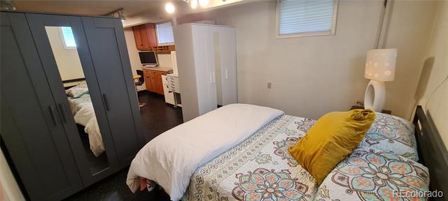 Furnished bedroom with patterned bedding, dark wardrobes, and a compact kitchen visible beyond.