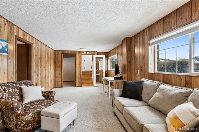 Living room with wood-paneled walls, a patterned armchair, and a sectional sofa beside a large window.