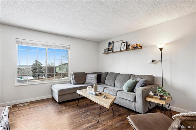 Living room with a gray sectional, wood-look floors, and a large window overlooking homes and trees.