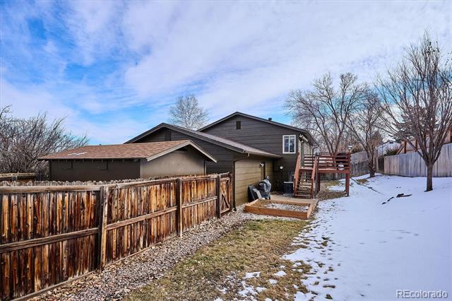 Rear of a dark-sided house with a wooden fenced yard, raised patio, and patchy snow.
