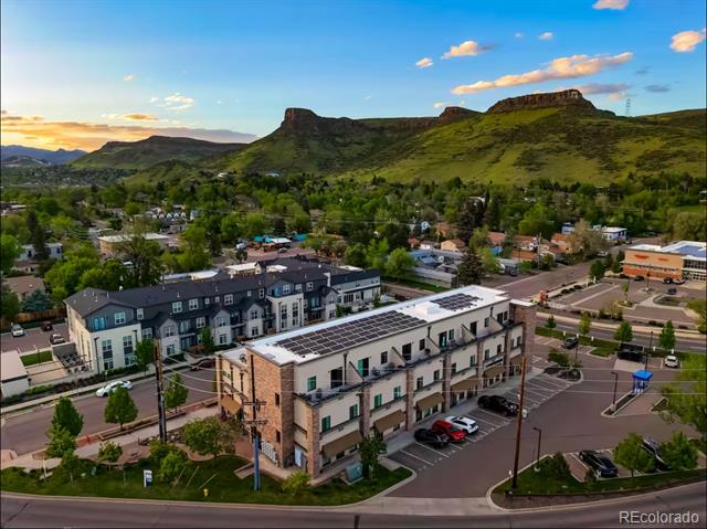 Modern residential building with rooftop solar panels, below green foothills and a flat-topped mountain.