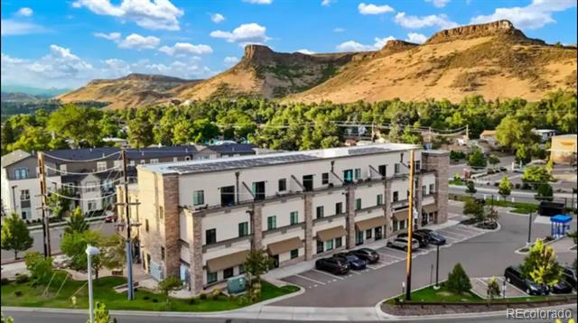 Multi-unit residential building with balconies and parking, backed by tree-covered foothills.