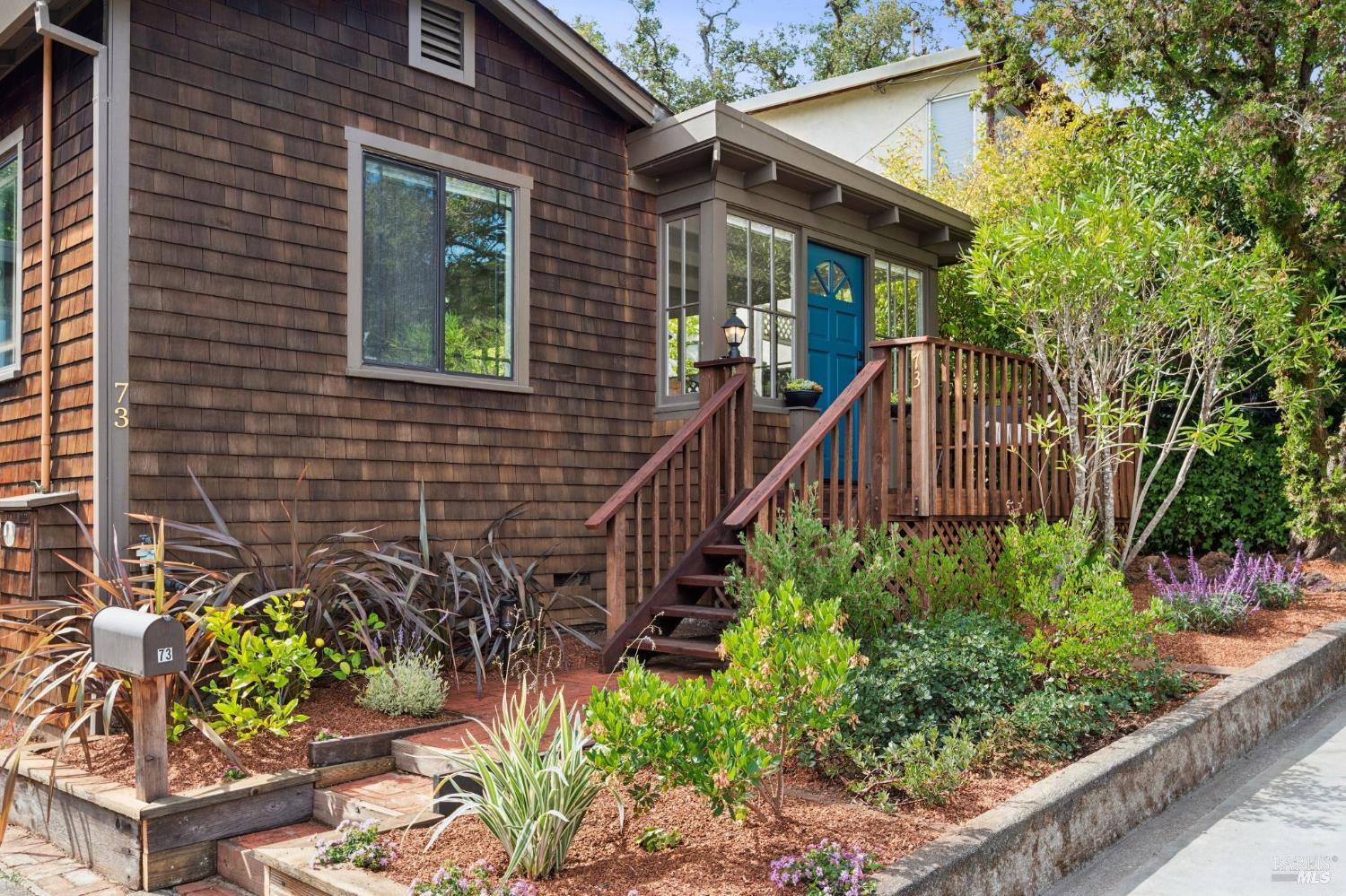 Two-story shingle home with a blue front door, wooden steps, and landscaped garden.