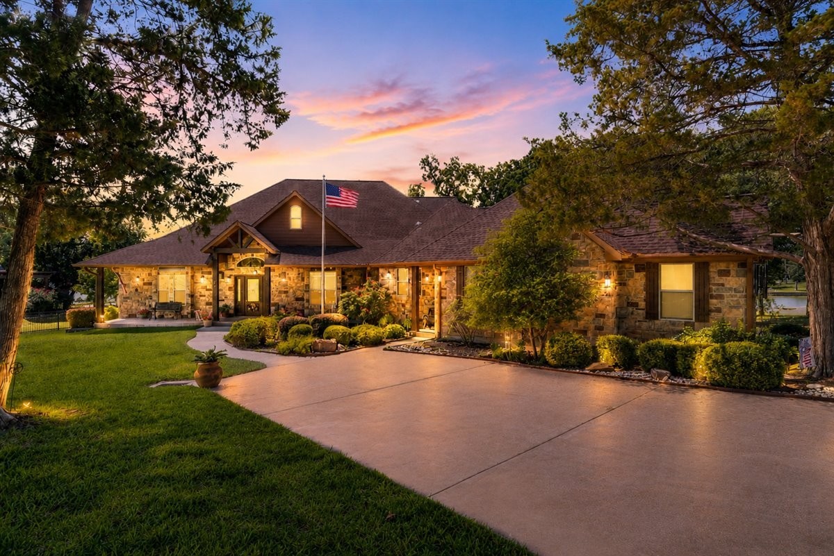 Stone home with an American flag and illuminated landscaping at dusk.