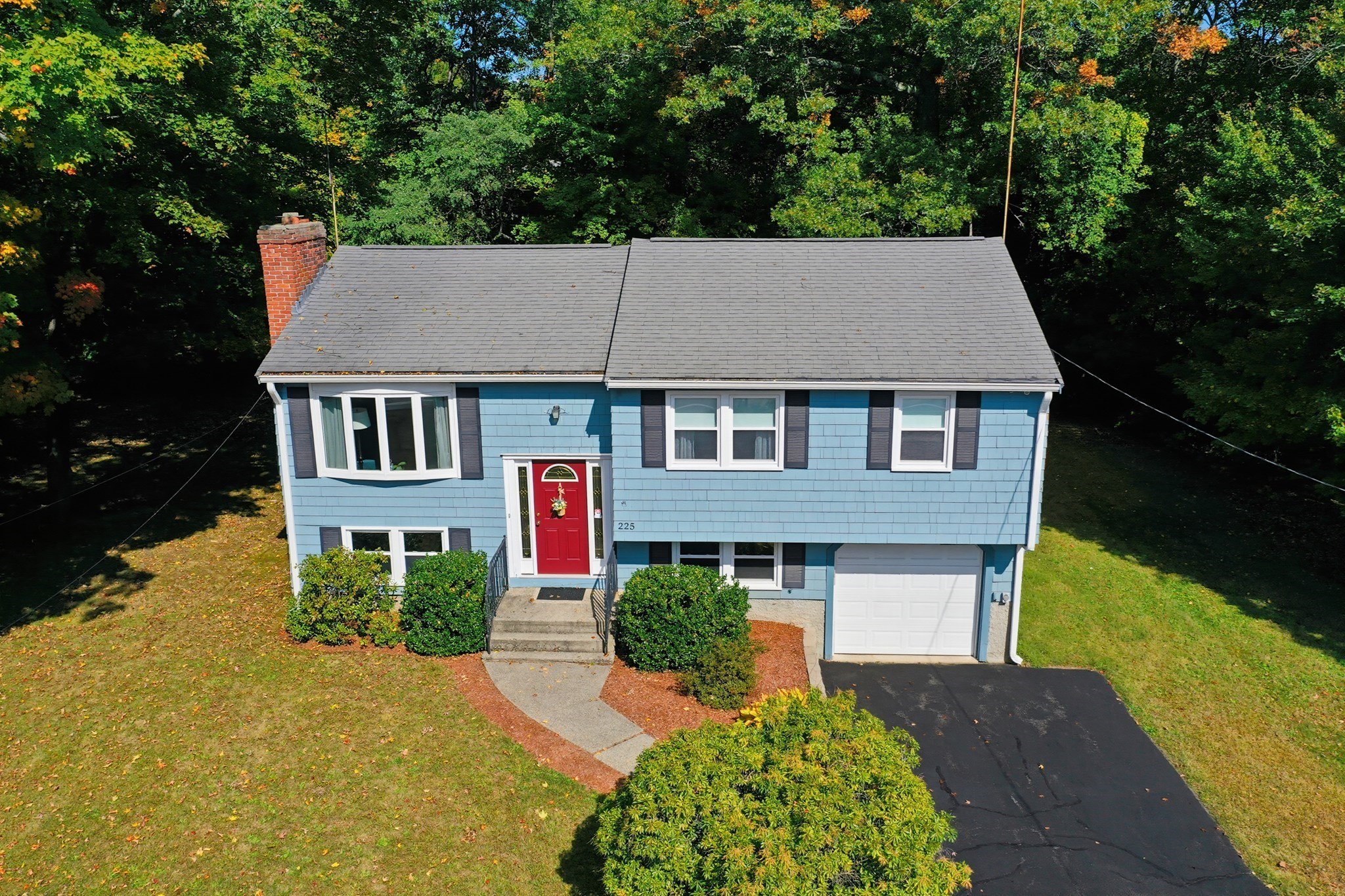 Aerial view of a blue split-level home with a red front door and attached garage, surrounded by trees.