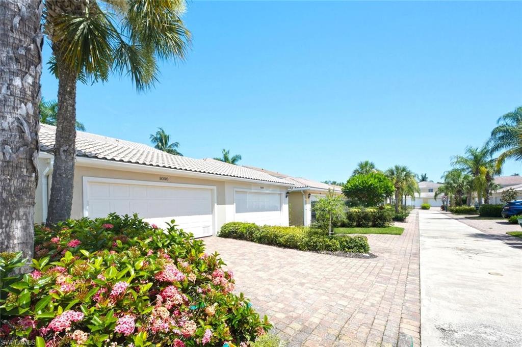 Home exterior with white garage doors, a brick-paver driveway, flowering shrubs, and palm trees.