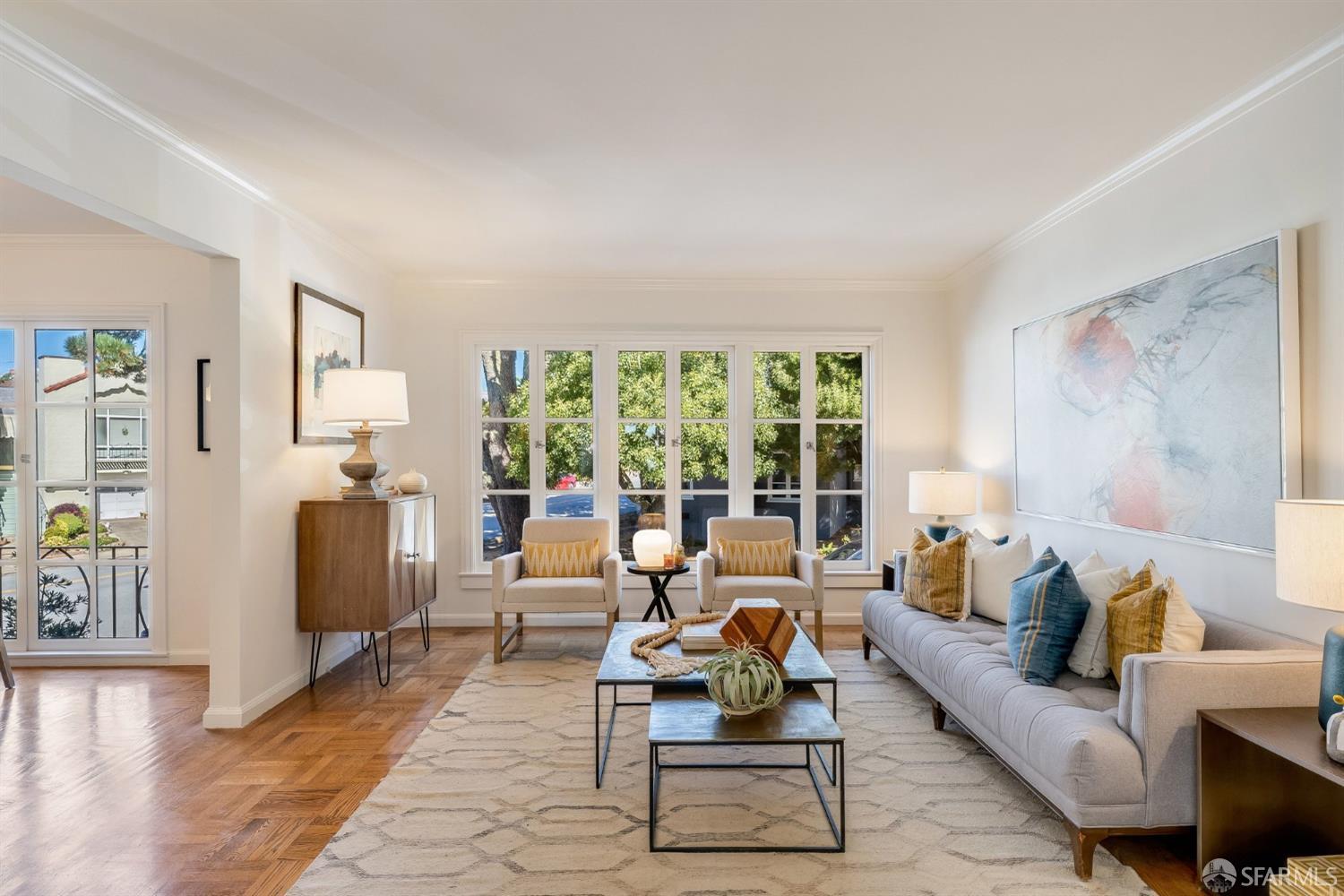 Furnished living room with a gray sofa, paired armchairs, and large windows overlooking trees.