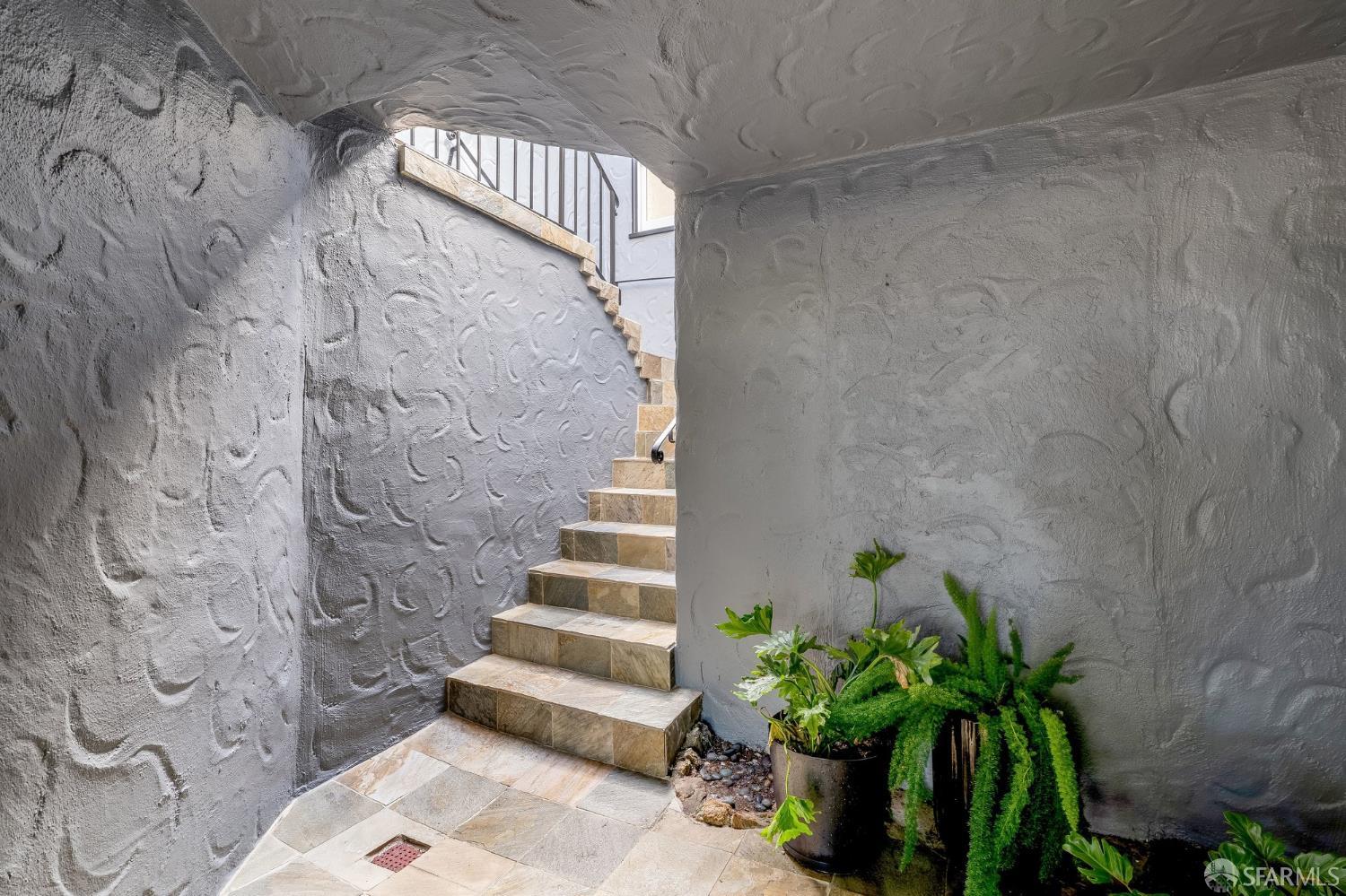 Tiled staircase rises between textured gray walls beside potted greenery.