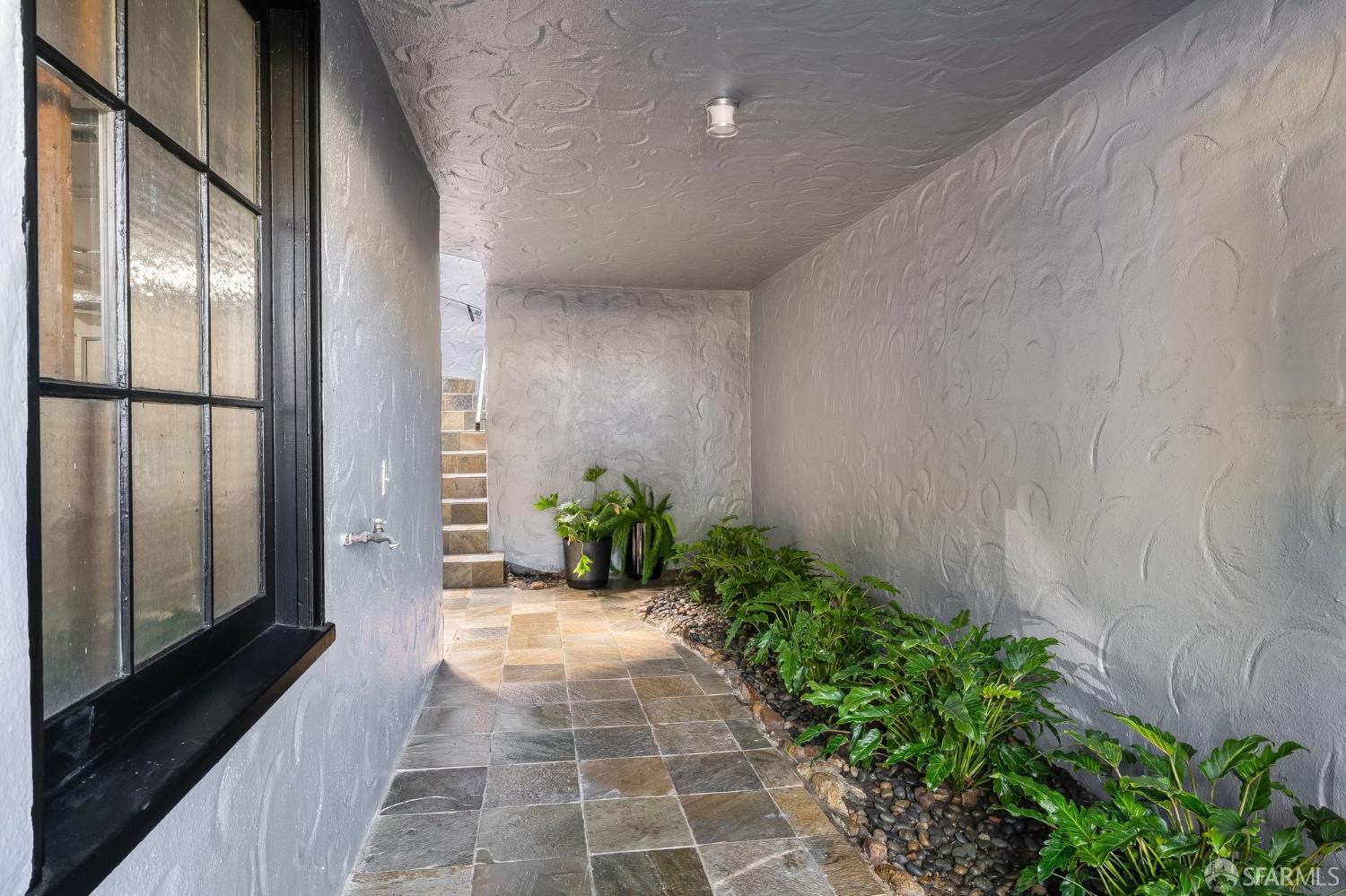 Covered entry walkway with textured stucco walls, stone tiles, leafy plants, and steps.
