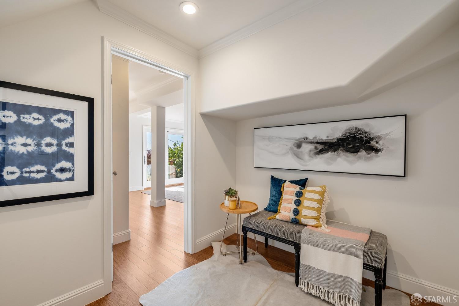 A staged sitting nook with a cushioned bench beneath abstract artwork, opening to a sunlit room.
