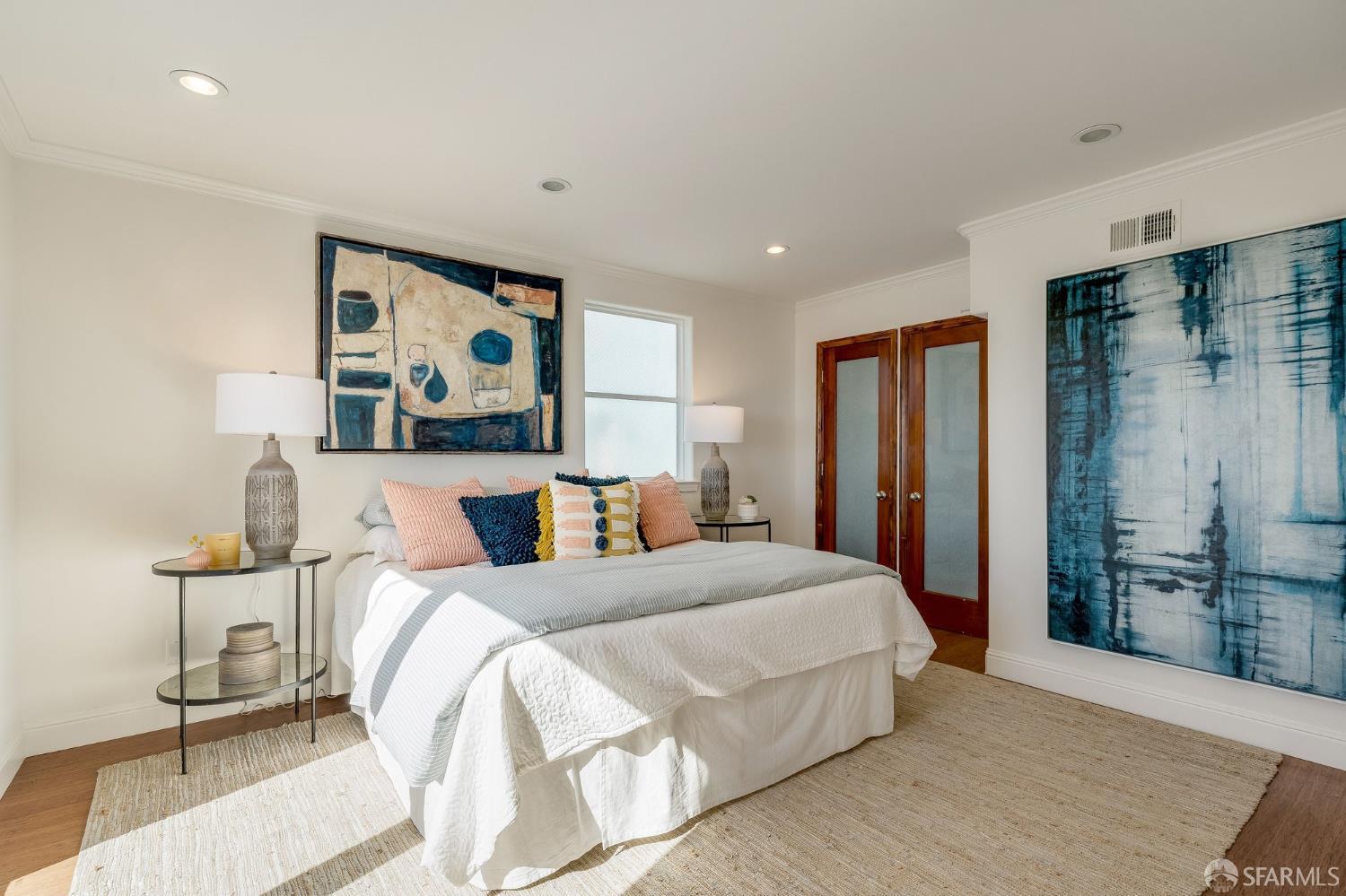 Staged bedroom with a white bed, colorful pillows, large abstract artwork, and sunlight across a neutral rug.