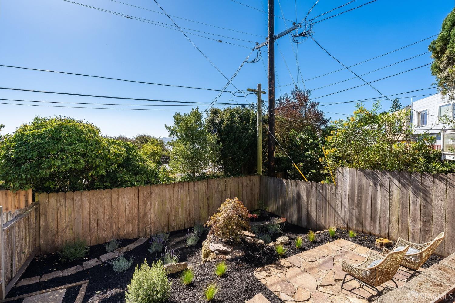 Backyard patio with flagstone paving, wicker chairs, garden beds, and a wooden privacy fence.