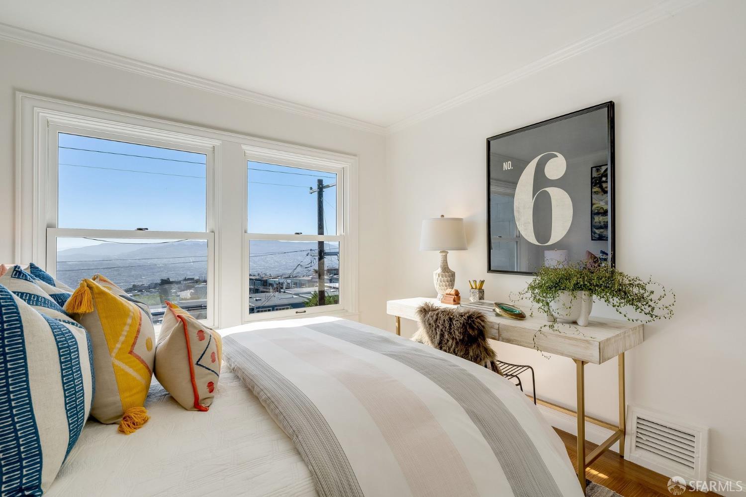 Staged bedroom with a white bed and colorful pillows, overlooking city rooftops and distant hills.