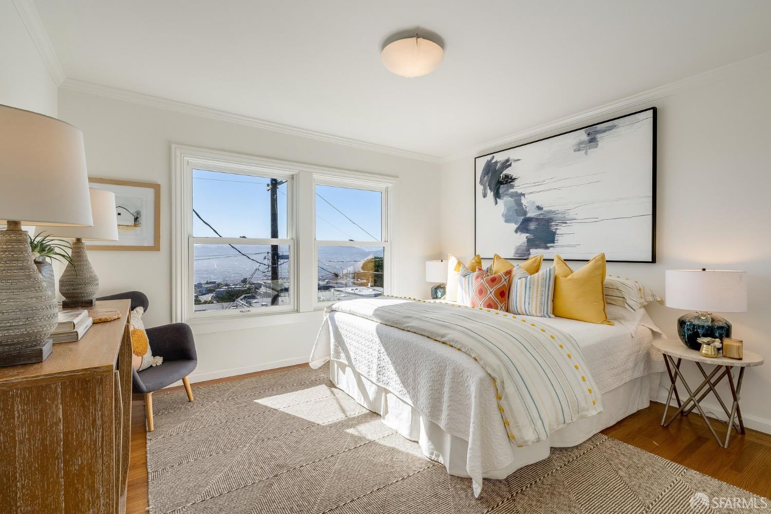 Bedroom with a white bed, yellow pillows, and city views through two windows.
