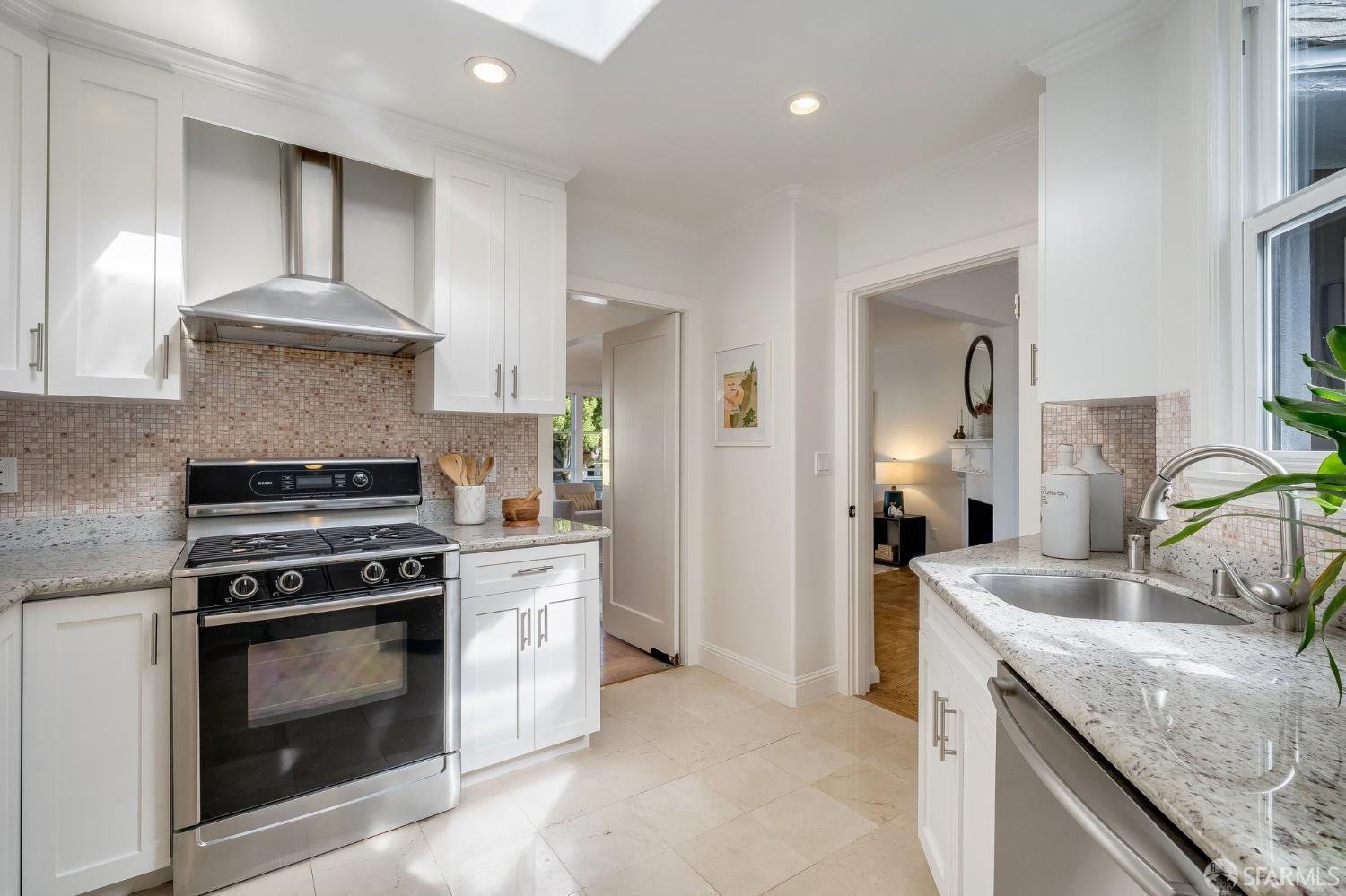 Bright white kitchen with a stainless-steel range, mosaic backsplash, and stone counters beside a large window.