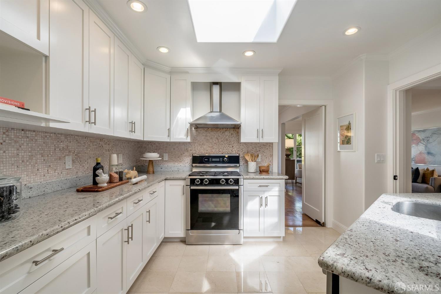Bright white kitchen with a skylight, tiled backsplash, stone counters, and stainless steel range.