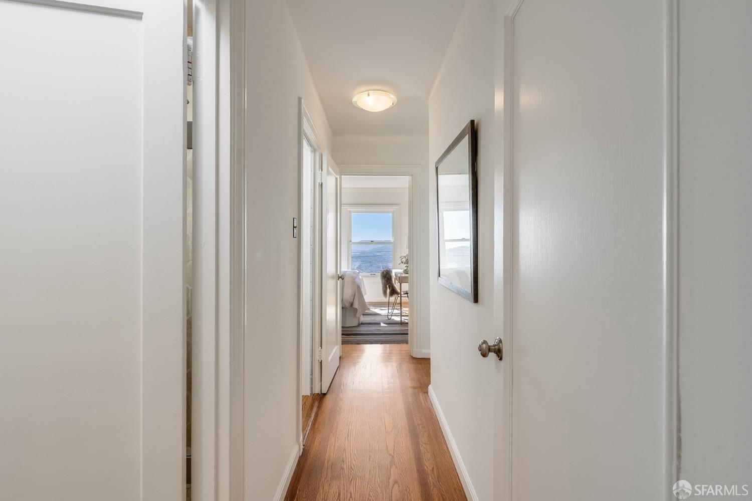 White hallway with hardwood floors leading to a bright furnished room and window view.