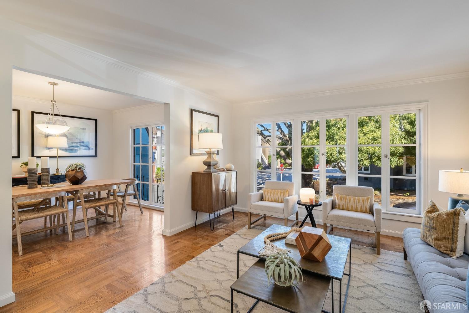 Staged living and dining room with hardwood floors and large windows overlooking leafy trees.