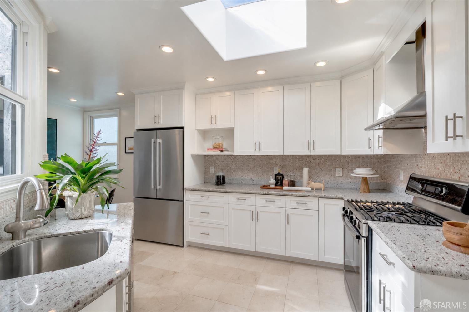 Bright white kitchen with a skylight, speckled stone counters, and stainless steel appliances.