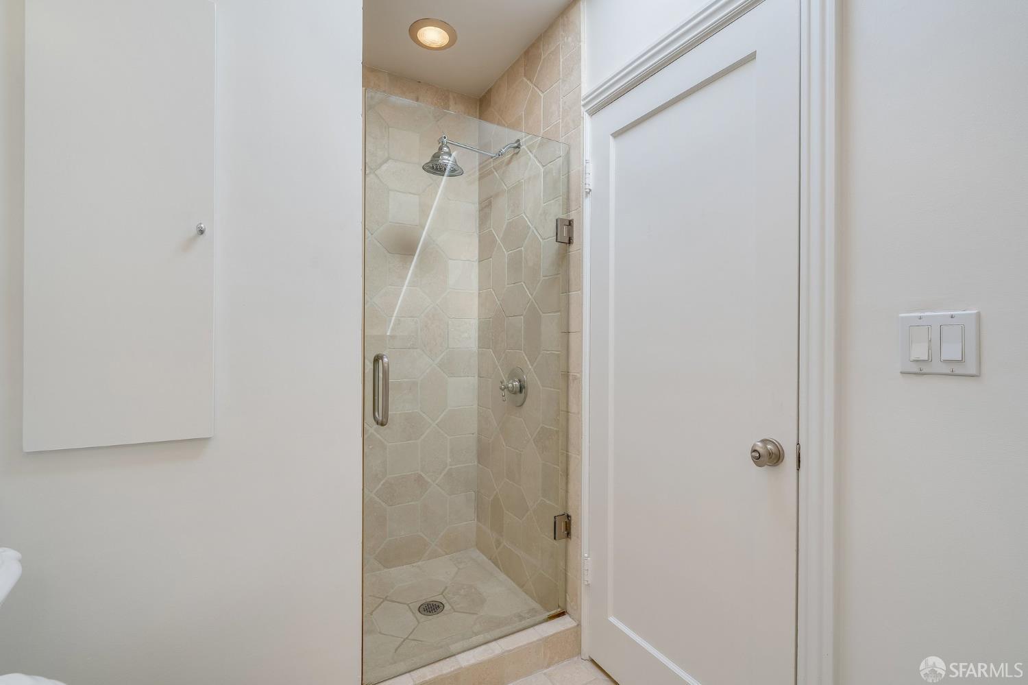 Walk-in shower with clear glass door, beige stone tile, and rainfall showerhead.