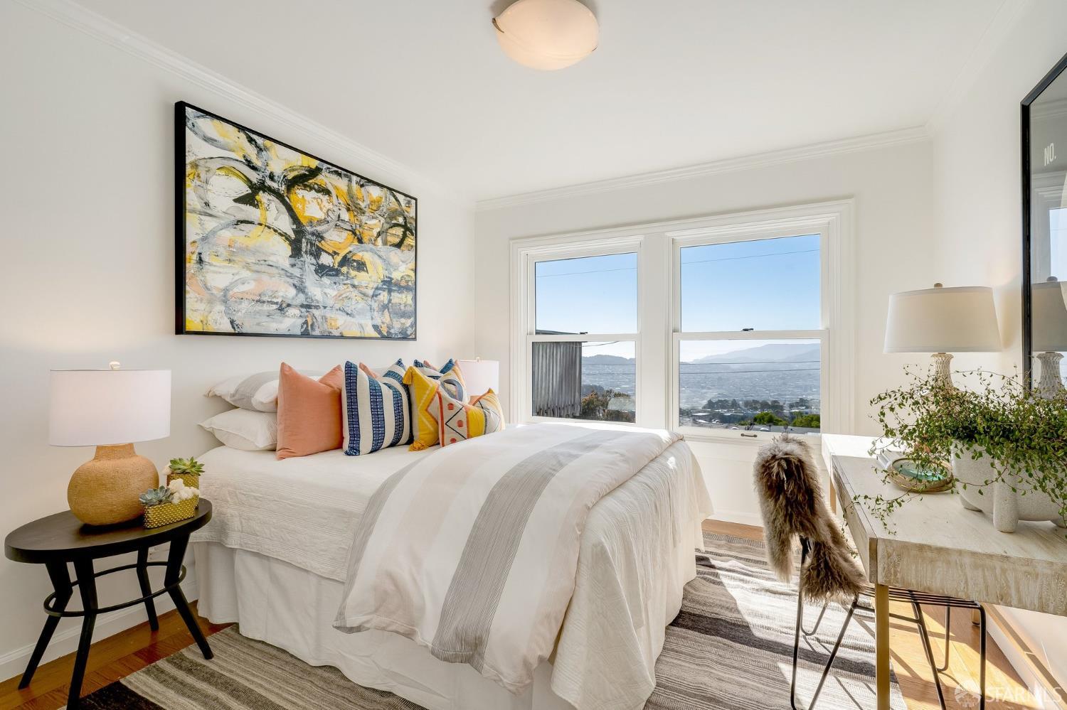 Staged bedroom with colorful pillows and large windows overlooking the city and distant hills.