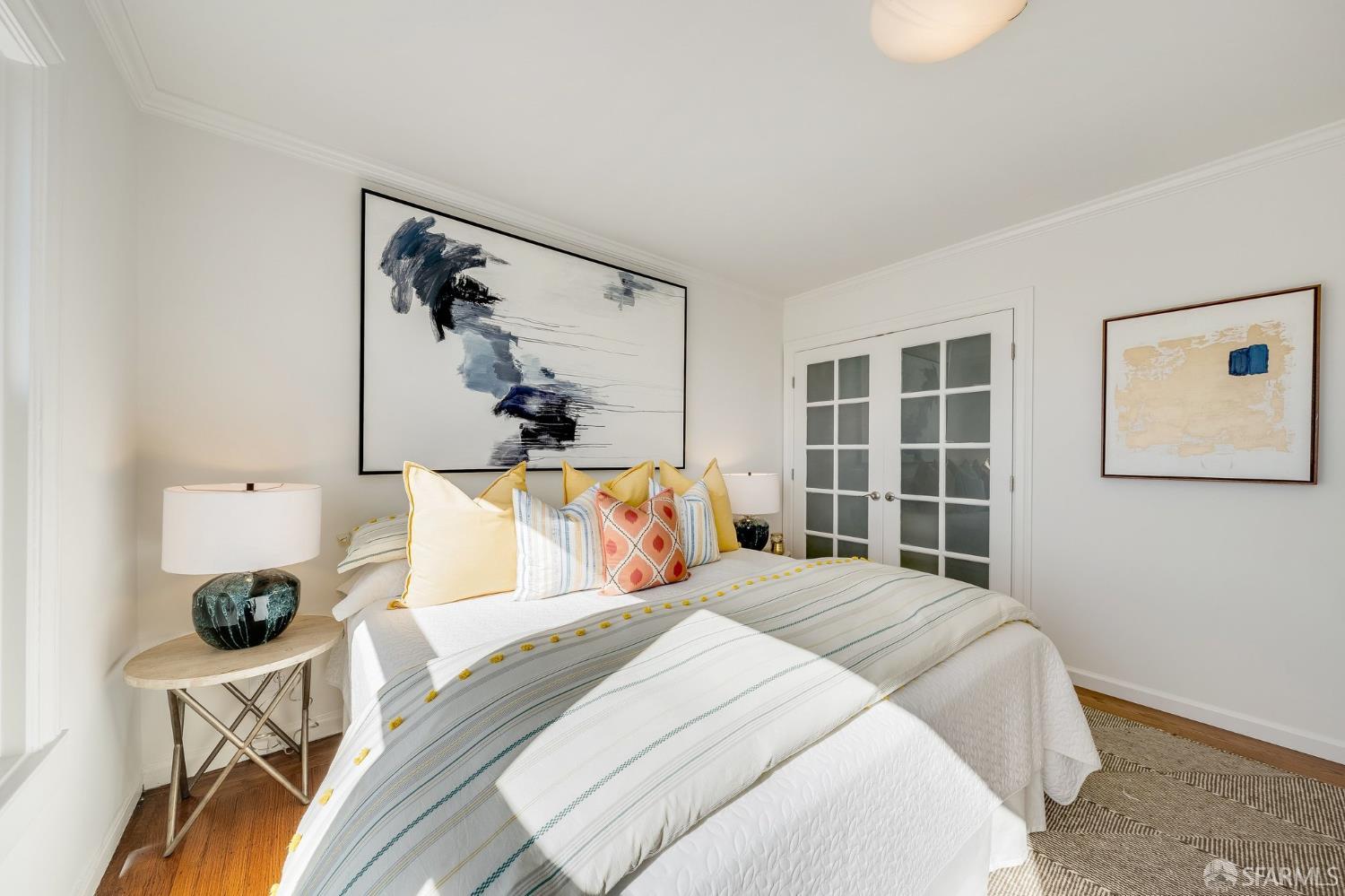 Staged bedroom with a white bed, colorful pillows, abstract artwork, and glass-paneled French doors.