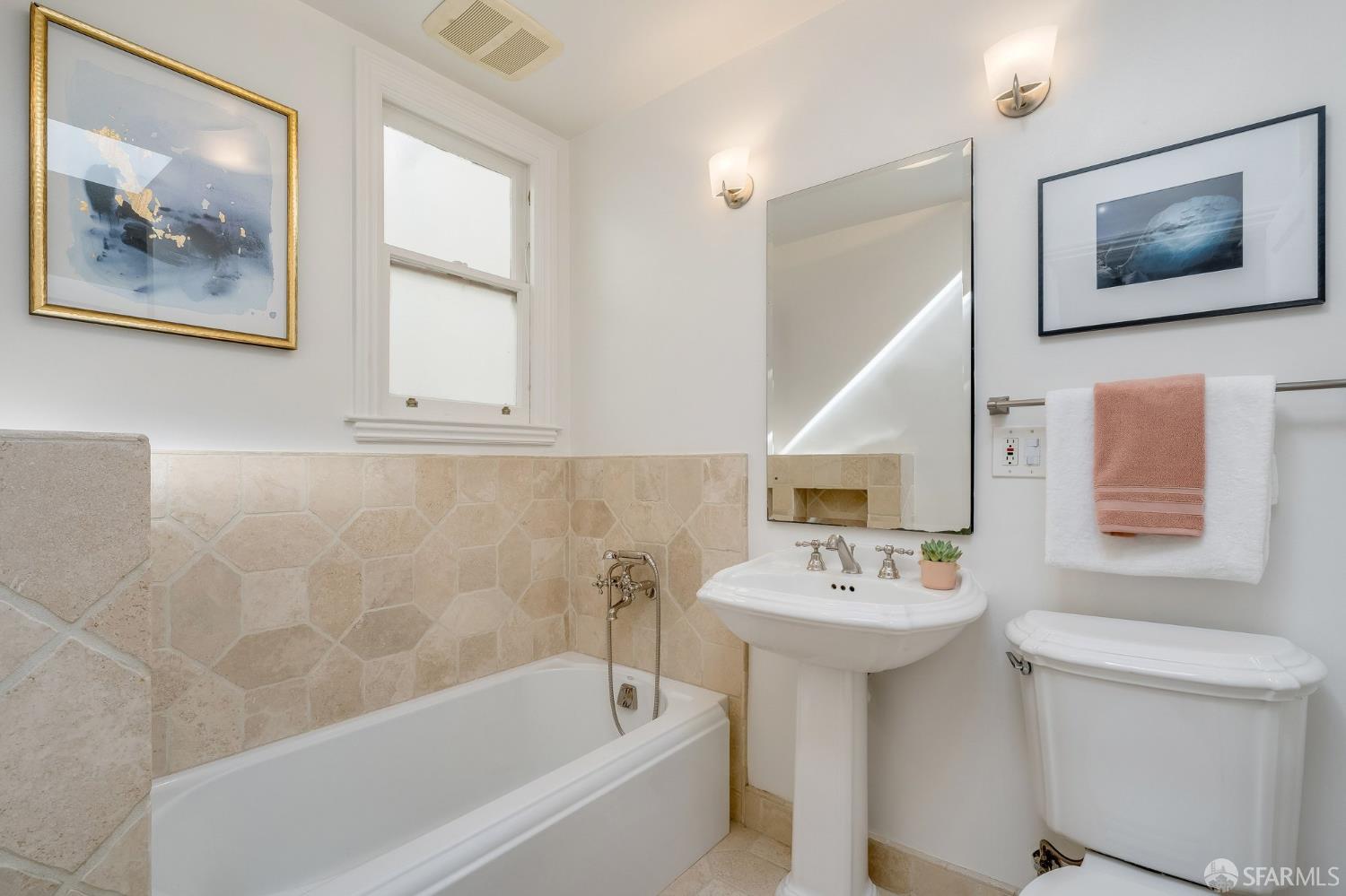 Bathroom with beige stone tile, a white bathtub, pedestal sink, and framed artwork.