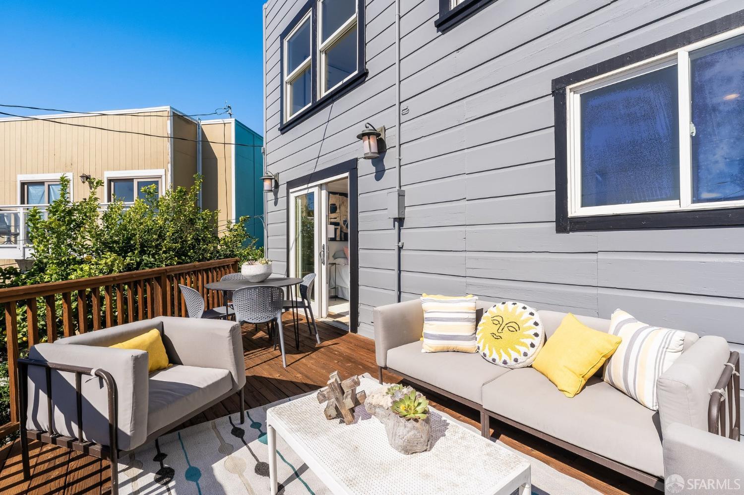 Furnished wooden deck with gray lounge seating, yellow pillows, and a dining table beside a gray-sided home.