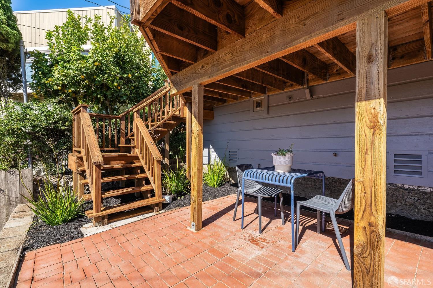 Covered brick patio with a wooden pergola, outdoor table and chairs, and garden stairs.