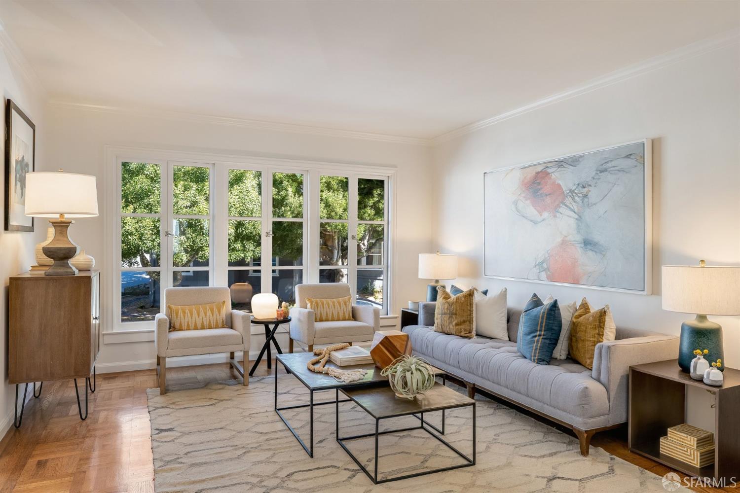 Staged living room with a gray sofa, cream armchairs, and large windows overlooking leafy trees.