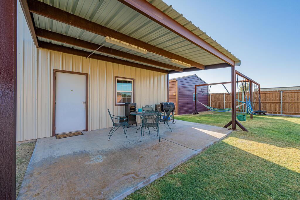 Covered patio with metal table and chairs beside a fenced grassy yard.