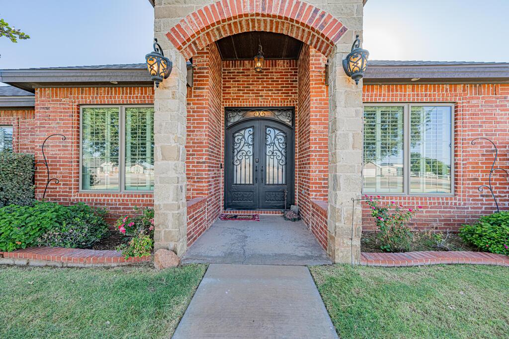 Red brick home entrance with a stone arch, ornate black double doors, and landscaped flower beds.
