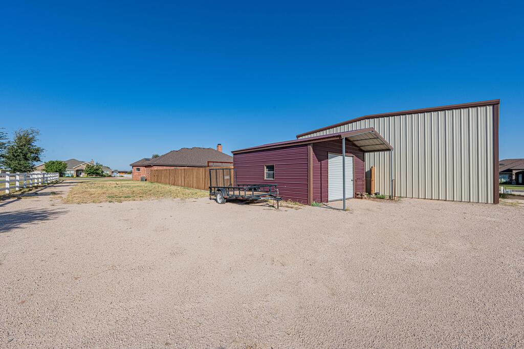 Maroon outbuilding beside a large metal workshop in a broad gravel yard.