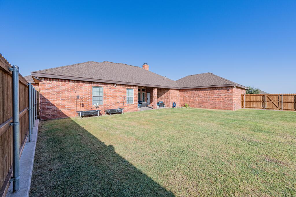 Single-story brick home behind a fenced lawn, with patio chairs along the rear wall.