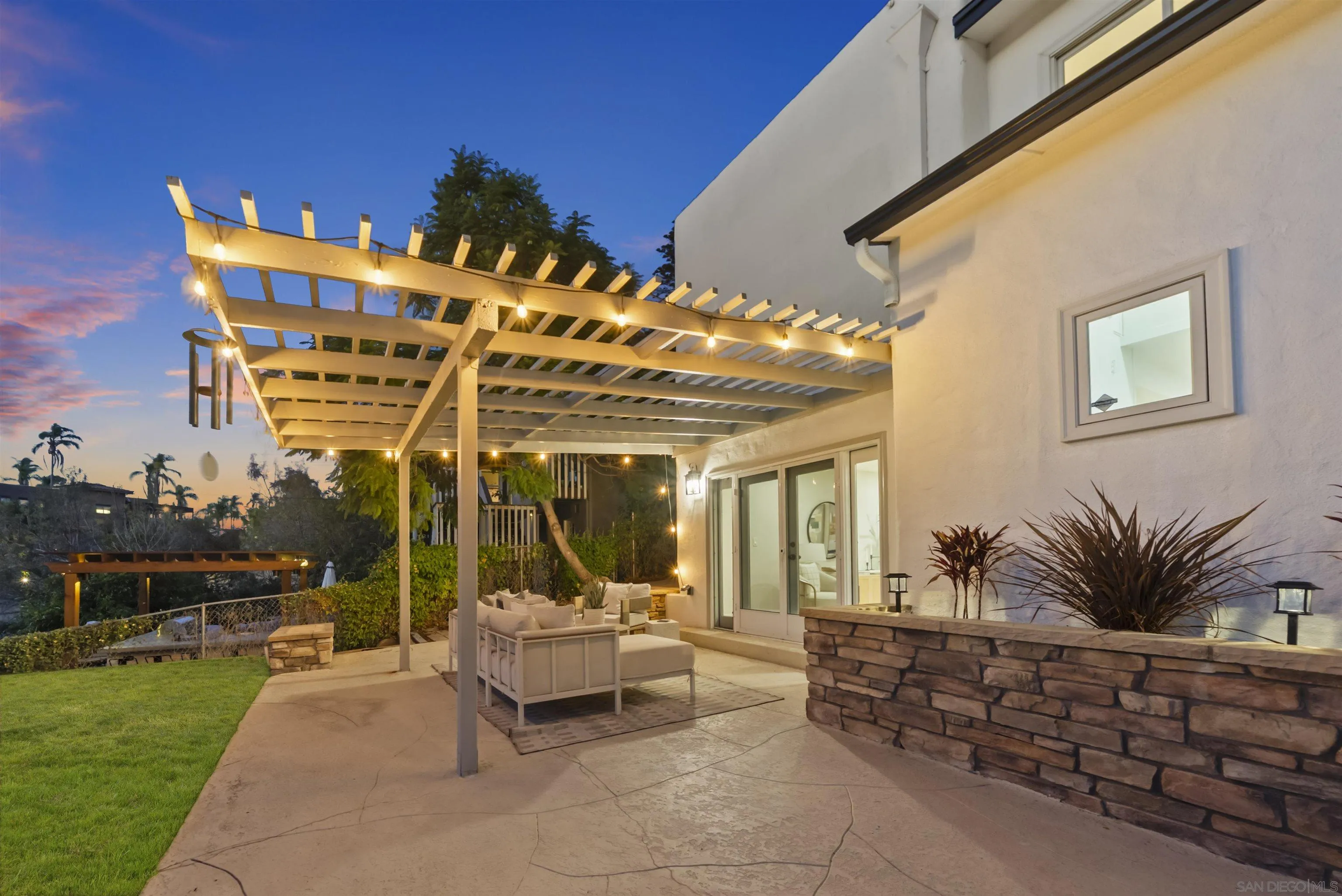 Backyard patio with a lighted white pergola, outdoor seating, lawn, and stone planter wall at dusk.