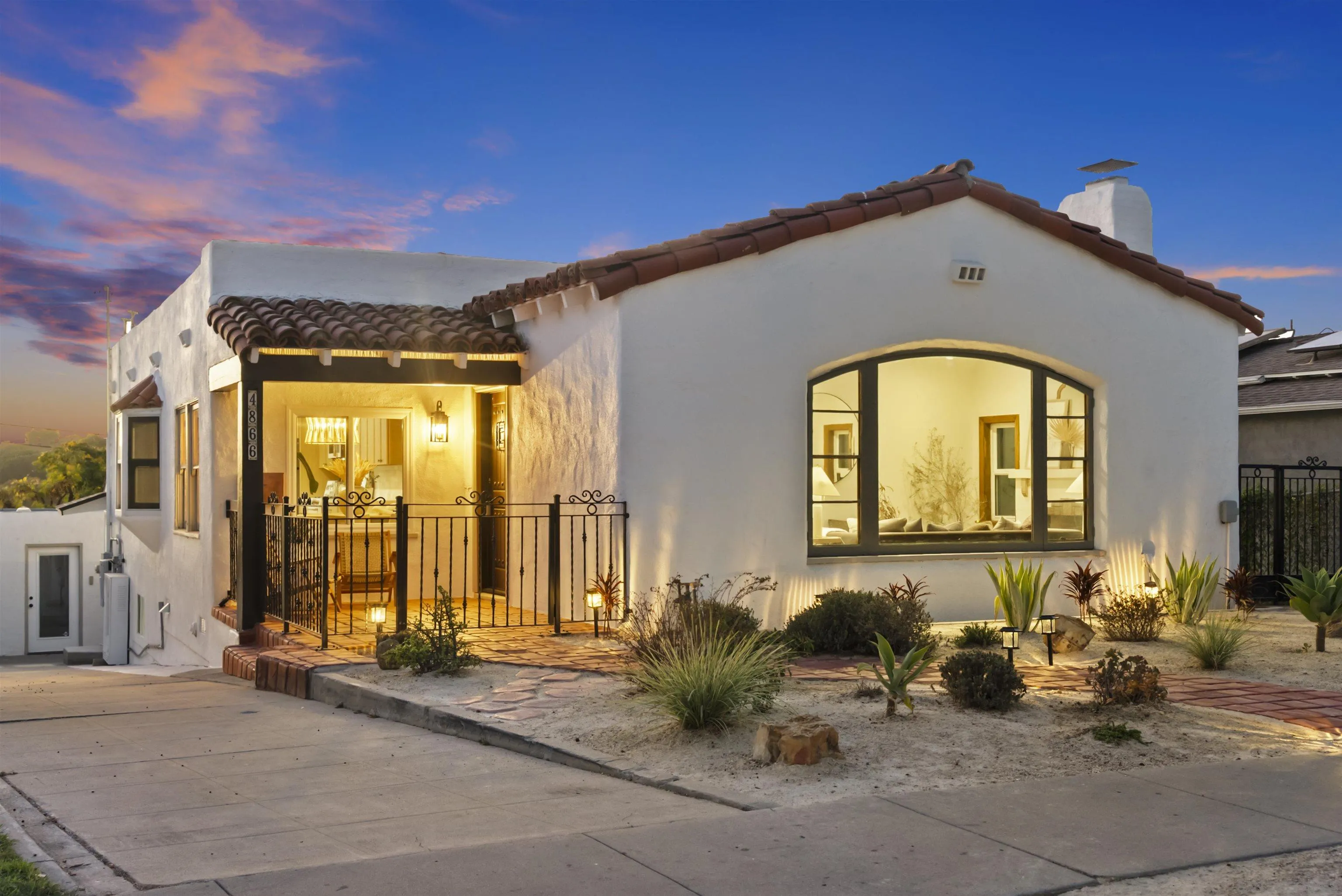 White stucco home with a red tile roof, arched window, and warmly lit entry at dusk.