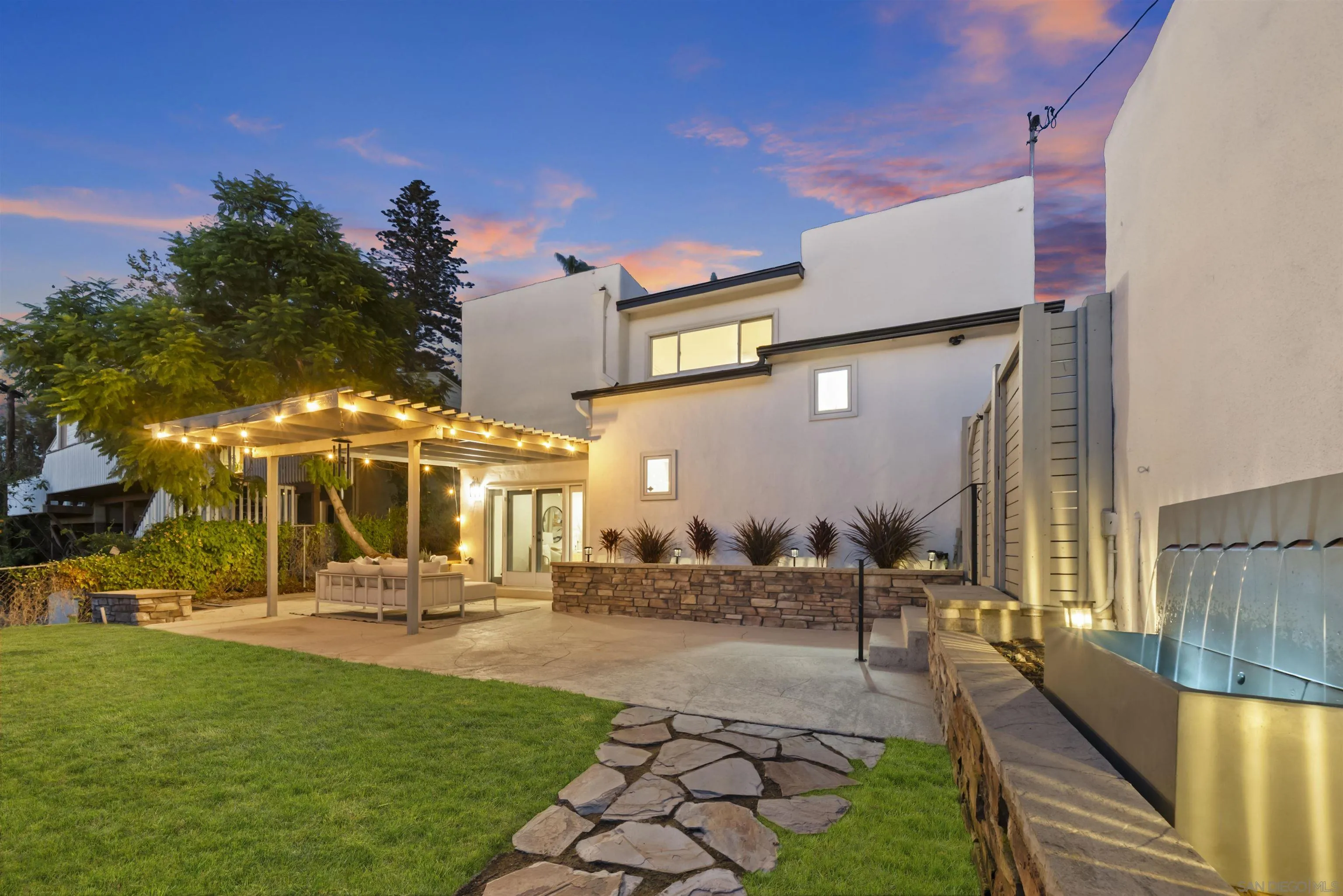 Modern home’s backyard at dusk, with a lighted pergola, stone path, and illuminated water feature.