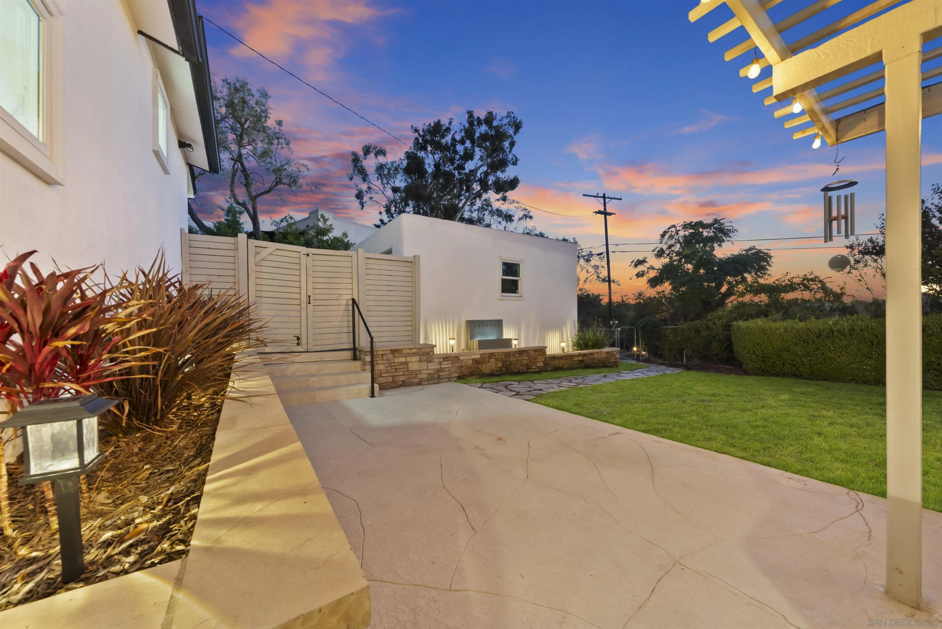 Backyard patio with a pergola, green lawn, and white outbuilding at sunset.
