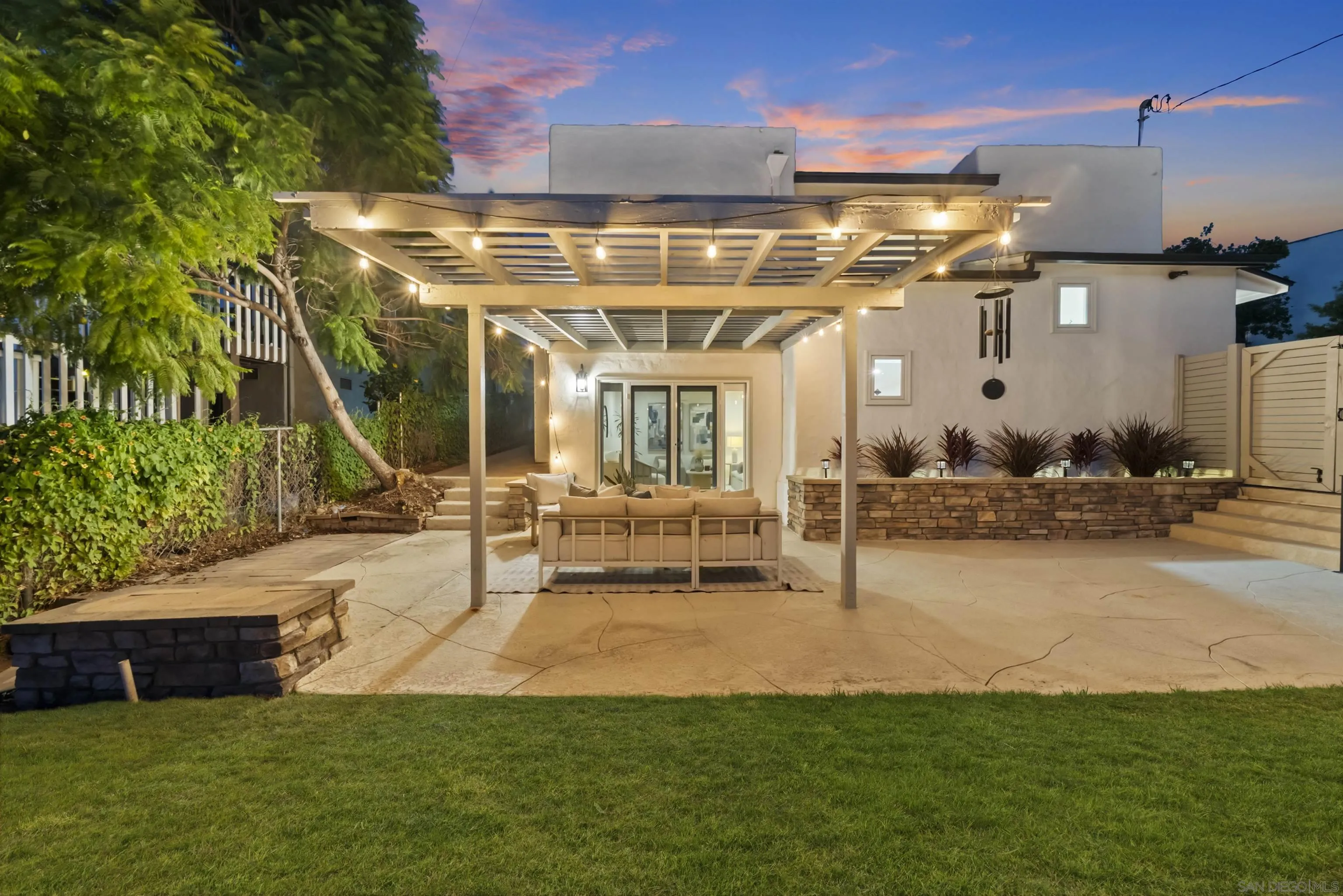 Backyard patio with a lighted pergola, outdoor seating, stone planter wall, and lawn at dusk.