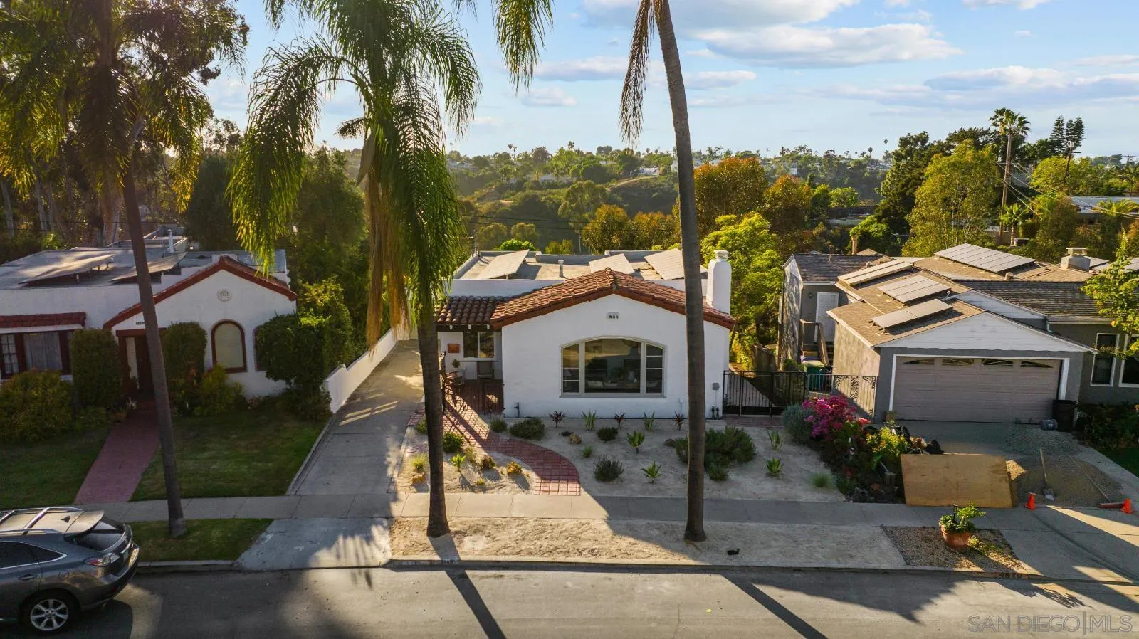 White single-story home with a red tile roof, framed by palm trees and overlooking green hills.