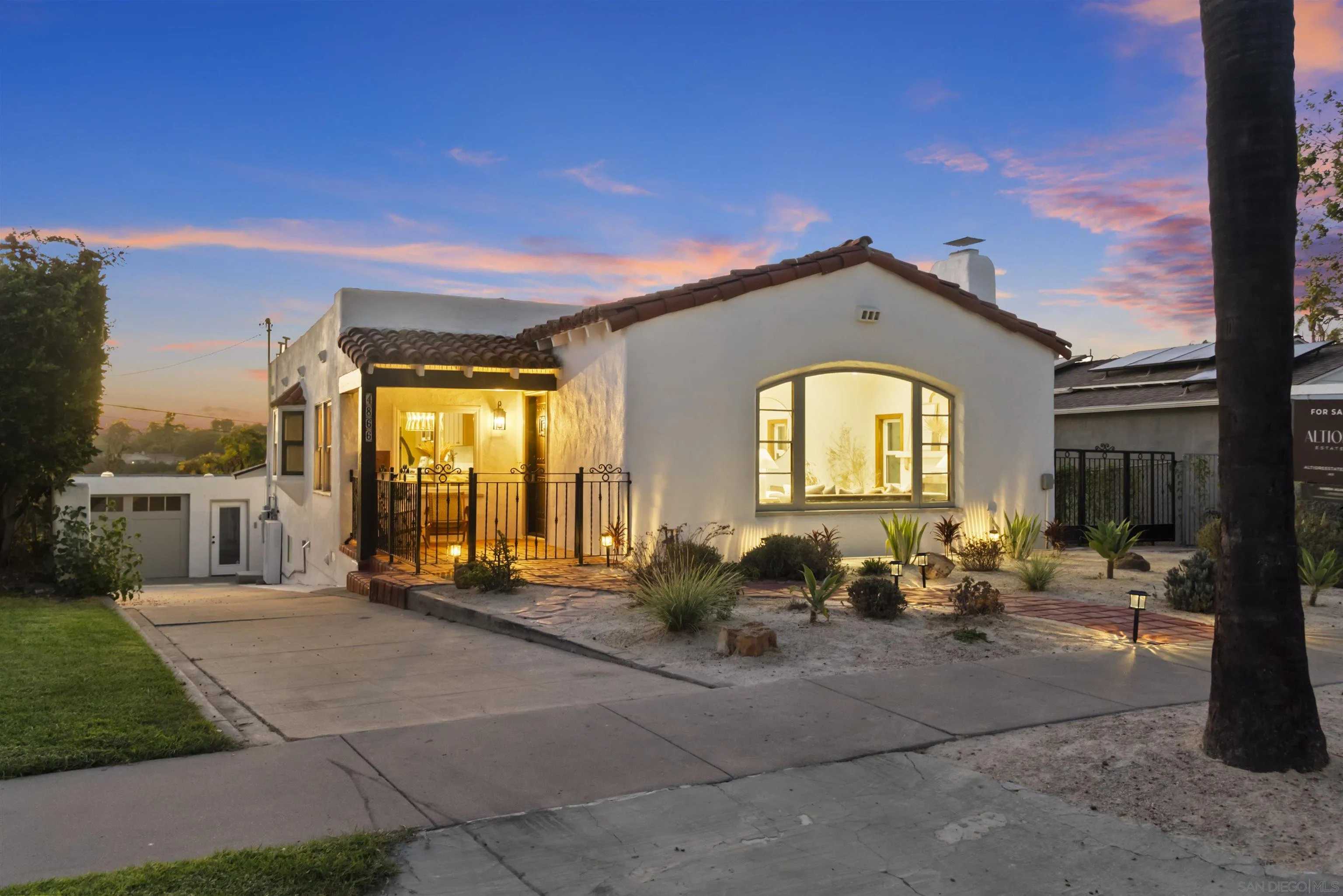White stucco home with terracotta roof, lit arched window, and landscaped front yard at dusk.
