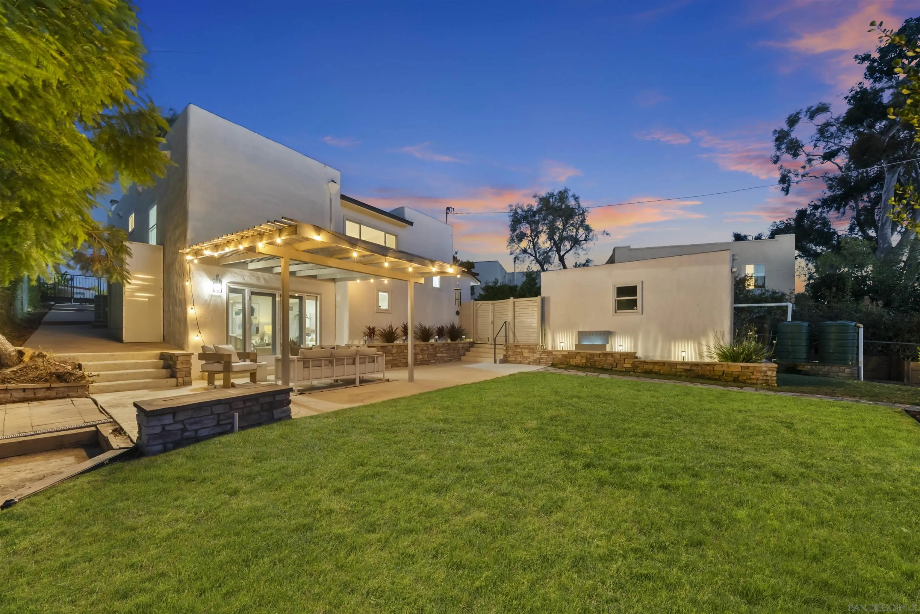 Modern home backyard with broad lawn and illuminated pergola at dusk.