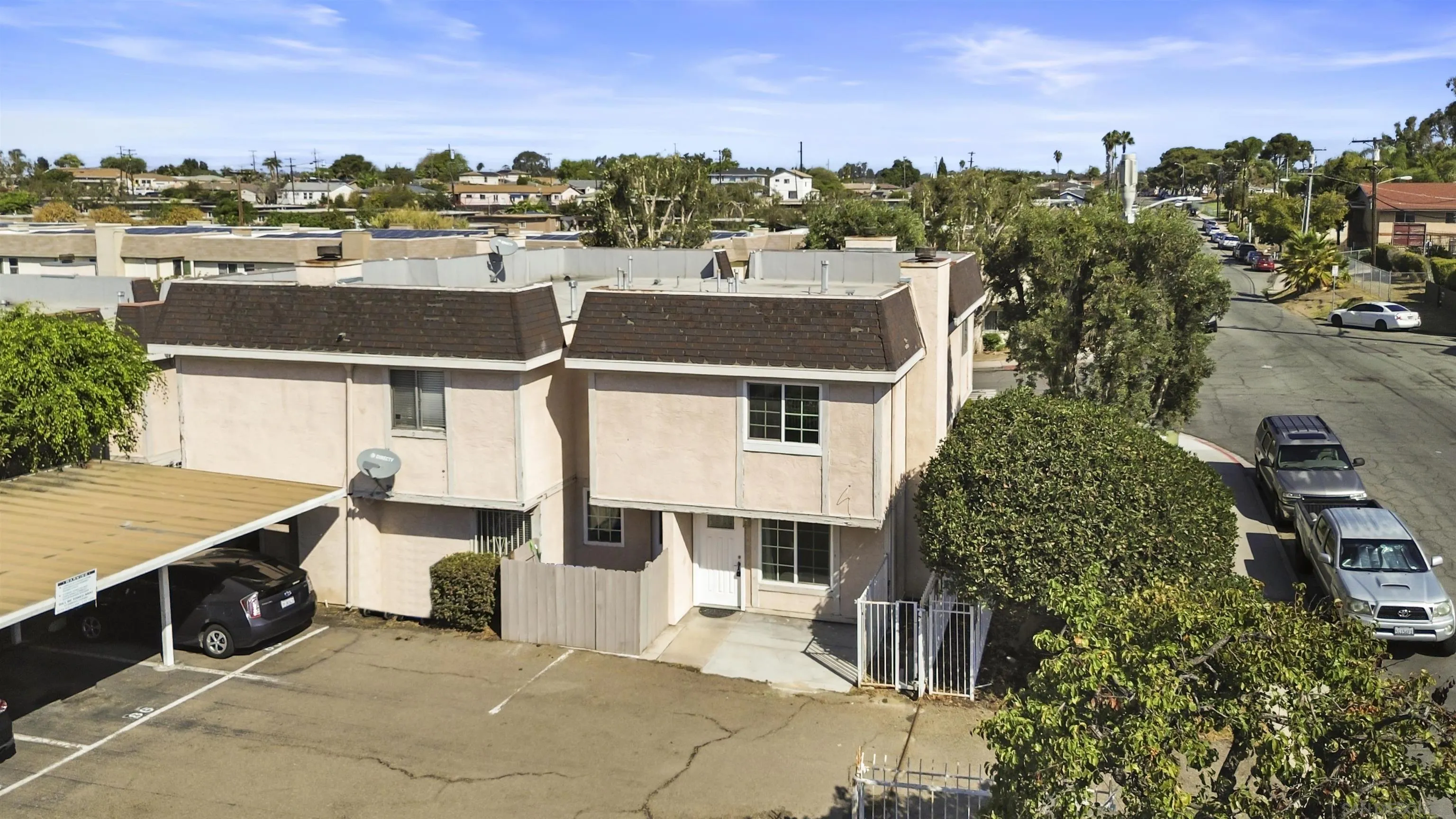 Two-story beige townhouse with a gated entry, large hedges, and adjacent parking.